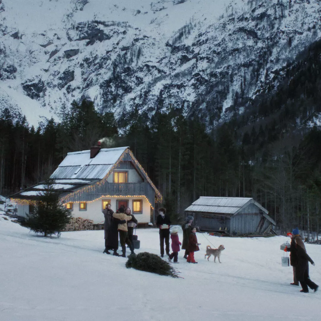People and a dog gather outside a house outlined with lights, with a felled tree in the snow and forested mountains behind.