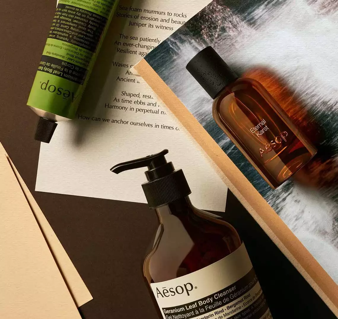 Aesop Eternal, Karst fragrance bottle, large amber pump bottle and cream tube on a dark surface with printed pages.